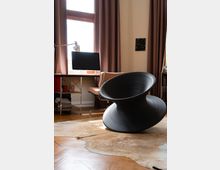 Helles Arbeitszimmer mit einem Schreibtisch, auf dem ein Computer und eine Lampe stehen, einem beige gepolsterten Bürostuhl und einem modernen, schwarzen drehbaren Stuhl in der Nähe. Der Raum hat braune Vorhänge, einen Holzboden mit einem beigefarbenen Teppich und ein großes Fenster, das viel Licht hereinlässt.