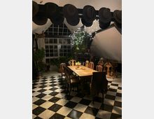 Großer Essbereich in einem loftartigen Raum mit schwarz-weiß kariertem Fliesenboden, langem Holztisch und gemischten Stühlen. Hohe Sprossenfenster, sichtbare Balken und eine Reihe von Hüten unter der Decke, dazu Pflanzen und Lichterketten als Dekoration.