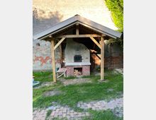 Backstein-Holzofen in einem Garten, überdacht von einer Holzkonstruktion mit Satteldach. Der Ofen steht vor einer gemauerten Wand, umgeben von Rasen und vereinzelten Pflastersteinen.