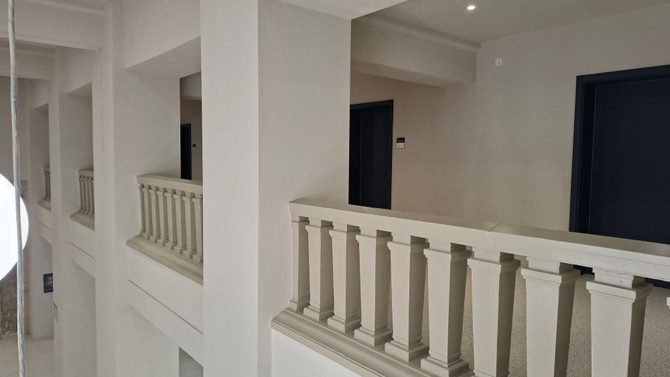Innenansicht eines hellen Flurs in einem mehrstöckigen Gebäude mit weißen Wänden, tragenden Säulen und Balustradengeländern. Entlang des Gangs befinden sich mehrere dunkle Türen, beleuchtet durch Deckenleuchten.