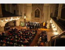 Innenraum einer Kirche mit hohen Wänden und gotischen Elementen. Ein Chor singt auf einer Bühne vor einem Altarbereich, während zahlreiche Besucher in Kirchenbänken sitzen; ein geschmückter Weihnachtsbaum steht links neben dem Chor.