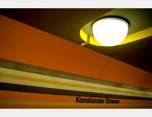 U-Bahn-Station mit orangefarbenen Wänden, einem gestreiften Mosaikband in Gelb-, Beige- und Brauntönen, der Aufschrift „Konstanzer Strasse“ und einer zentralen Deckenleuchte mit abgerundetem Design.