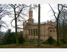 Backsteinkirche im romanischen Stil, umgeben von kahlen Bäumen in einer waldähnlichen Umgebung. Das Gebäude hat hohe Rundbogenfenster, einen Turm mit grünlicher Kuppel und detaillierte Fassadenelemente.