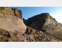 Sandige Klippenformationen unter klarem blauem Himmel, mit einzelnen Gräsern auf den oberen Kanten. Die erodierten Schichten zeigen unterschiedliche Erd- und Sandfarben.