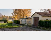 Gelände mit einem einfachen Gebäude und Garage, umgeben von Zäunen und Hecken; im Hintergrund ein Baum mit Herbstlaub und ein Blick auf einen See in einer ländlichen Umgebung. Im Vordergrund befinden sich Grünflächen, Sitzbänke und ein Gehweg.