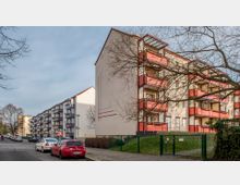 Mehrfamilienhäuser mit roten Balkonen in einem Wohngebiet, flankiert von parkenden Autos entlang einer ruhigen Straße. Im Vordergrund ist ein großer Baum ohne Laub und ein Zaun mit einem kleinen Tor sichtbar.