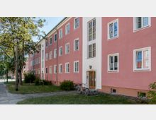 Mehrstöckiges Wohngebäude mit rosafarbenen Fassaden und weißen Fensterrahmen. Vor dem Gebäude befinden sich Grünflächen, Bäume, ein Gehweg und ein Fahrradständer mit zwei abgestellten Fahrrädern.