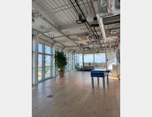 Open-plan modern office lounge with floor-to-ceiling windows, wood flooring, and an exposed industrial ceiling with ducts, pipes, and hanging lights. A foosball table, large potted plant, and seating area sit near glass doors leading to a terrace with a city view.