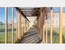Ein gläserner, rechteckiger Korridor mit Holzbalken und Backsteinpfosten, der von Kletterpflanzen bewachsen ist, führt über einen Kiesweg. Im Hintergrund sind eine Wiese und Gebäude mit großen Fenstern sichtbar.