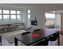 Helles Wohnzimmer mit großen Fenstern und Blick auf eine Stadtlandschaft, ausgestattet mit einem Esstisch, Hängeleuchten, einem Sessel und einem Wandfernseher; daneben ein Zugang zu einem Balkon mit Liegestühlen. Ein Bücherregal erstreckt sich unter den Fenstern.