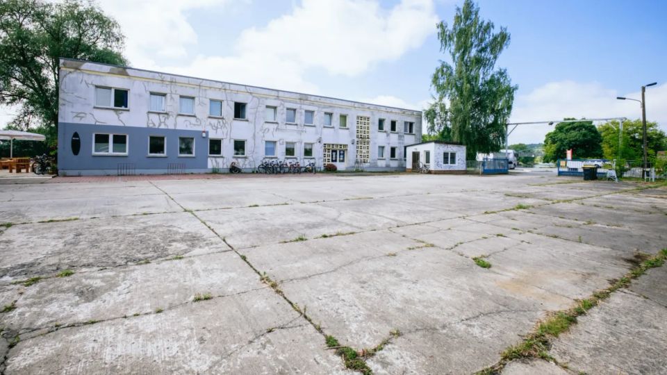 Ein zweistöckiges Gebäude mit einer schlichten weißen Fassade und rechteckigen Fenstern steht hinter einer großen, gepflasterten Fläche. Im Vordergrund sind vereinzelt Fahrräder abgestellt, und im Hintergrund sind Bäume und Zaunstrukturen sichtbar.