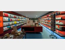 Innenansicht eines modernen Buchladens mit roten Regalen, die vielfältige Bücher und Publikationen ausstellen. Ein zentraler Tisch zeigt weitere Auslagen, und ein rotierender Ständer rechts enthält Postkarten. Der Raum hat einen blauen Boden und helle Deckenbeleuchtung.