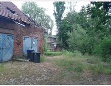 Ein altes, teilweise verfallenes Backsteingebäude mit blauen Holztoren, umgeben von Gras, Bäumen und dichtem Grün. Im Hintergrund ist ein weiteres Gebäude zu sehen, eingebettet in eine bewaldete Umgebung.