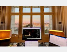 Blick aus einem Büro mit großen Fenstern auf eine Stadtlandschaft bei Sonnenuntergang, mit Gebäuden, Bäumen und einem entfernten Fernsehturm. Im Vordergrund stehen zwei Monitore auf einem Schreibtisch, umgeben von Lampen und Büromaterialien.