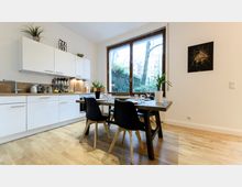 Moderne Küche mit weißer Einbauküche, Holzarbeitsplatte und Hängeschränken, daneben ein Esstisch mit dunkler Holzplatte und vier schwarzen Stühlen. Im Hintergrund ein großes Fenster mit Blick ins Grüne und Dekorationen wie Zimmerpflanzen und Wandbild.