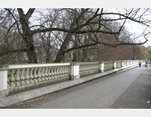 Gehweg entlang einer historischen Balustrade aus weißem Stein, flankiert von kahlen Bäumen in einer urbanen Parkumgebung. Im Hintergrund sind Personen zu Fuß unterwegs.
