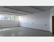 Ein leerer, rechteckiger Raum mit weißen Wänden und einer Decke mit Rasterplatten und Leuchtstofflampen. Auf der linken Wand befinden sich große Fenster, die Tageslicht hereinlassen, und darunter ist ein Heizkörper angebracht.