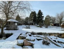Verschneiter Garten mit einer kleinen Holzhütte, einem Steg und einem angrenzenden Teich. Im Hintergrund sind Bäume, Büsche, ein Holzzaun und weitere Gebäude zu sehen.