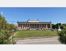 Neoklassizistisches Gebäude mit einer breiten Säulenfront (Altes Museum in Berlin) vor einer großen Rasenfläche und einem Brunnen; im Hintergrund weht eine deutsche Flagge, umgeben von Bäumen und einem klaren blauen Himmel.