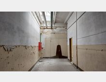 Ein verlassener Flur mit stark abgenutzten Wänden, an denen die Farbe abblättert. An der Decke verlaufen freiliegende Rohre, und ein roter Feuerlöschkasten ist an der Wand angebracht; der Boden ist mit Staub und Schutt bedeckt.