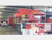 Innenansicht einer Feuerwehrgarage mit mehreren geparkten roten Einsatzfahrzeugen, darunter eine Drehleiter und ein Anhänger. Im Vordergrund befinden sich technische Geräte wie eine Wasserpumpe, während an der Decke Belüftungsrohre und Halterungen für die Abgasschläuche sichtbar sind.
