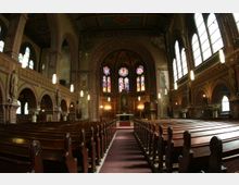 Innenraum einer historischen Kirche mit roten Ziegelwänden, Rundbögen und Holzbankreihen, die zum Altar führen. Über dem Altar befinden sich bunte Glasfenster mit religiösen Motiven, flankiert von Wandmalereien und Statuen.