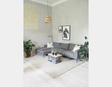 Modernes Wohnzimmer mit hellgrauen Wänden, einem grauen Ecksofa mit weißen Kissen, einem kleinen Holztisch mit Metallgestell und einem gewebten Teppich. Die Einrichtung wird durch grüne Zimmerpflanzen, zwei minimalistische Wandbilder und eine Hängelampe aus geflochtenem Material ergänzt.