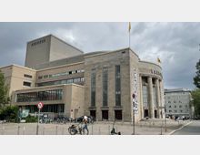 Außenansicht eines monumentalen Theaters im neoklassizistischen Stil mit einer halbkreisförmigen Fassade, flankiert von Säulen und hohen Fenstern. Im Vordergrund befindet sich ein Platz mit Fahrrädern, E-Scootern und einer Person, und im Hintergrund ist ein bewölkter Himmel sichtbar.