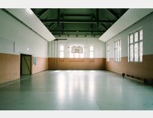 Innenraum einer einfachen Turnhalle mit hohen Decken und sichtbaren Dachträgern. Der Raum hat eine Basketballkorbanlage, große Fenster mit Gitterstruktur und helle Wände, die teilweise mit Holzpaneelen verkleidet sind.