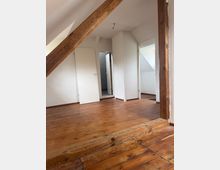 Leerer Dachgeschossraum mit schrägen Decken, sichtbaren Holzbalken und breiten Holzdielen. Weiße Wände mit mehreren offenen Türen führen in angrenzende kleine Räume.