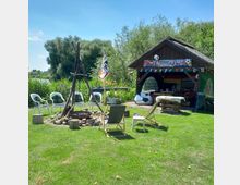 Grüne Wiese mit einer Feuerstelle aus Steinen, umgeben von Gartenstühlen und Holzhockern; im Hintergrund ein überdachter Pavillon mit Fußball-Dekorationen und Wimpelgirlanden, nahe einem Fluss und umgeben von dichtem Grün.