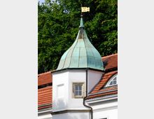 Weißer Gebäudeturm mit grün oxidierter Kupferkuppel und vergoldeter Wetterfahne, flankiert von einem roten Ziegeldach; im Hintergrund sind Bäume zu sehen.