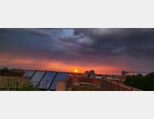 Sonnenuntergang über einer Stadtlandschaft mit ziegelgedeckten Dächern, Solarpanels auf einem Gebäudevorsprung und einem Geländer im Vordergrund. Der Himmel ist von orangefarbenen und violetten Wolken durchzogen, während im Hintergrund Baukräne und hohe Gebäude sichtbar sind.