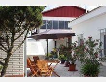 Hofbereich mit Holztisch und passenden Stühlen unter einem großen grauen Sonnenschirm, umgeben von Pflanzen in Töpfen und einem Baum links im Vordergrund. Im Hintergrund sind weiße und rote Gebäudewände mit Fenstern und Solarpanels sichtbar.