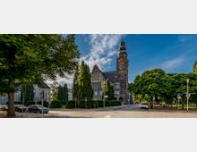Historisches Gebäude mit Turm und großer Uhr, umgeben von Bäumen und gepflegten Hecken, an einer gepflasterten Straße mit geparkten Autos und Fußwegen. Der Himmel ist klar mit einigen Wolken.