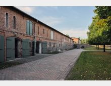 Langes, einstöckiges Gebäude aus Backstein mit grünen Holztüren und Steinmauersockel, flankiert von einer gepflasterten Einfahrt und einer Grünfläche mit Bäumen. Der Himmel ist leicht bewölkt, und im Hintergrund sind geparkte Autos zu sehen.