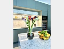 Moderne Küche mit einer grau-marmorierten Arbeitsplatte, auf der eine Vase mit bunten Calla-Lilien und ein Korb mit Zitrusfrüchten (Zitronen und Limetten) stehen. Im Hintergrund befinden sich grüne Schränke, eine Einbau-Weinkühlung, sowie offene Regale mit Flaschen.