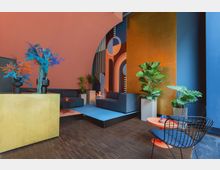 Moderner Innenraum mit lebhaften Farben, darunter eine Wand mit geometrischen Mustern in Blau, Orange und Gelb. Der Raum enthält stilvolle Möbel wie blaue Sofas, schwarze Drahtstühle und bunte Beistelltische sowie zahlreiche Pflanzen und auffällige Trockenblumenarrangements.