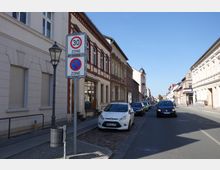 Straßenansicht einer kleinen Stadt mit zwei- bis dreistöckigen Häusern, meistens in hellen Putz- und Backsteinfassaden. Im Vordergrund stehen Verkehrsschilder für eine Tempo-30-Zone und ein Parkverbot, während Autos entlang der Straße geparkt sind; im Hintergrund ist eine Kirche mit Zwiebelturm sichtbar.