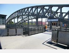 Eine metallene Bahnbrücke mit stahlverstärkten Bögen und Trägern, darunter ein Bahnsteig mit Geländern und einer Wegweisung zur Station "Bornholmer Straße". Im Hintergrund sind bunte Wohnhäuser und grüne Bäume zu sehen.