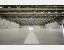 Große, leere Industrie- oder Messehalle mit glatten Betonwänden und offener Decke, an der zahlreiche Leitungen und Lampen befestigt sind. Der Boden ist weitgehend frei, mit einem schmalen Streifen in der Mitte, der in Richtung einer verglasten Eingangstür verläuft.