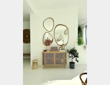 Minimalistisches Wohnzimmer mit einem Holzschrank mit Lamellenfronten, dekoriert mit Blumen, Vasen und Büchern. An der weißen Wand hängen drei verschieden geformte Spiegel mit Holzrahmen, daneben steht eine Topfpflanze.