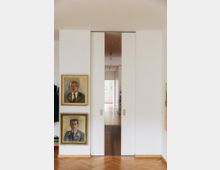 Moderner Innenraum mit weiß gestrichenen Wänden und Schiebetüren, die in einen Flur mit Holzboden führen. Links an der Wand hängen zwei gerahmte Porträtgemälde übereinander, und der Boden zeigt ein Fischgrätparkett.