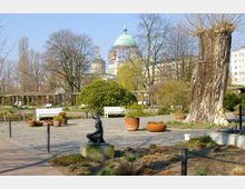 Ein gepflegter Park mit Sitzbänken, Pflanzkübeln und einer kleinen Bronze-Skulptur im Vordergrund, umgeben von Bäumen und Sträuchern, im Hintergrund ist eine Kuppel eines historischen Gebäudes sichtbar.