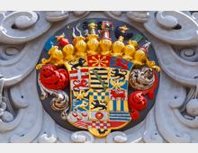 Wappen mit detailreich gestalteten Symbolen und goldener Krone, umgeben von kunstvollen grauen Verzierungen; zeigt verschiedene farbige Felder mit Löwen, Kreuzen, Streifen und anderen heraldischen Elementen.