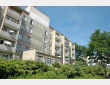 Wohnblock aus Betonplatten mit fünf Stockwerken und Balkonen, umgeben von dichter grüner Vegetation und einzelnen Bäumen unter einem klaren blauen Himmel.