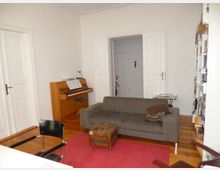 Wohnzimmer mit Holzboden, grauem Sofa, rotem Teppich und einem Holzhocker. Im Hintergrund ein Klavier mit Notenblättern, eine offene Tür mit weißen Flügeltüren, sowie ein Bücherregal auf der rechten Seite.