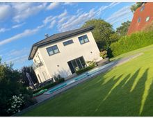 Modernes, zweigeschossiges Wohnhaus mit heller Fassade und dunklem Satteldach an einem großen, gepflegten Rasen. Im Vordergrund liegt ein rechteckiger Pool mit umlaufender Terrasse, umgeben von Hecken und Bäumen unter blauem Himmel.
