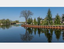 Flusslandschaft mit klarem Wasser und einer befestigten Uferpromenade aus rostfarbenen Metallplatten; im Hintergrund Bäume, Büsche und vereinzelte Gebäude unter blauem Himmel.
