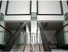 Zwei parallel verlaufende Rolltreppen führen in einem modern gestalteten, hell beleuchteten Innenraum nach oben. Die Wände sind weiß, mit vereinzelten schwarzen Akzentelementen und quadratischen, grünlichen Lichtkästen.